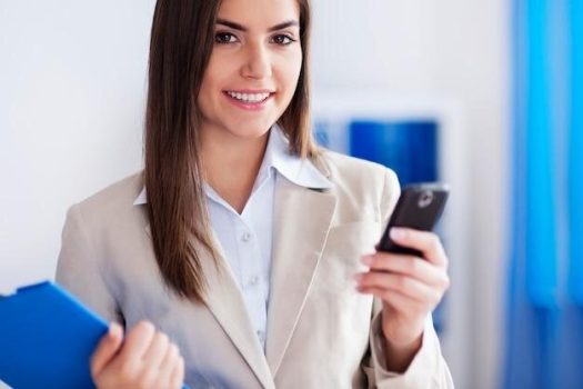 Mulher sorridente em escritório usa o celular, representando o app para corretoras de seguros e o atendimento digital