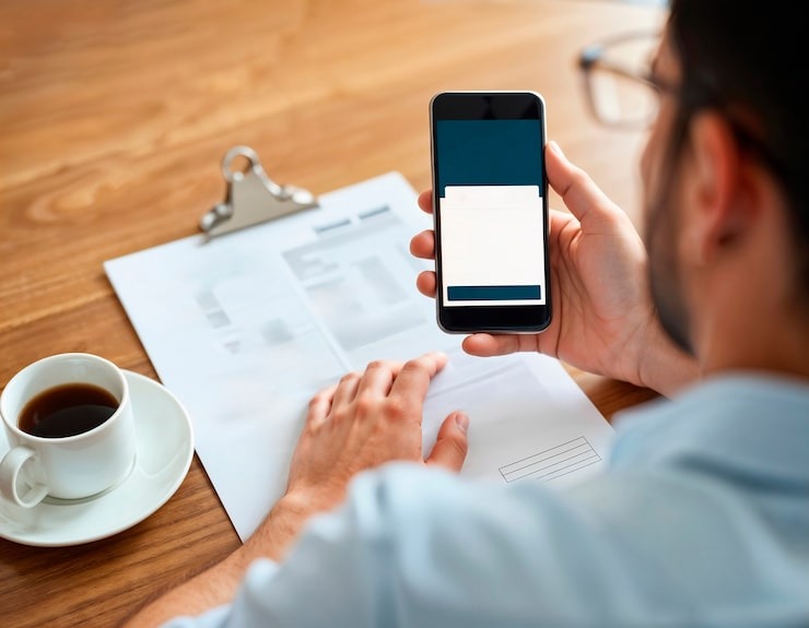 Homem consulta apólice no celular sobre documentos, ilustrando app para corretoras de seguros com a marca da corretora